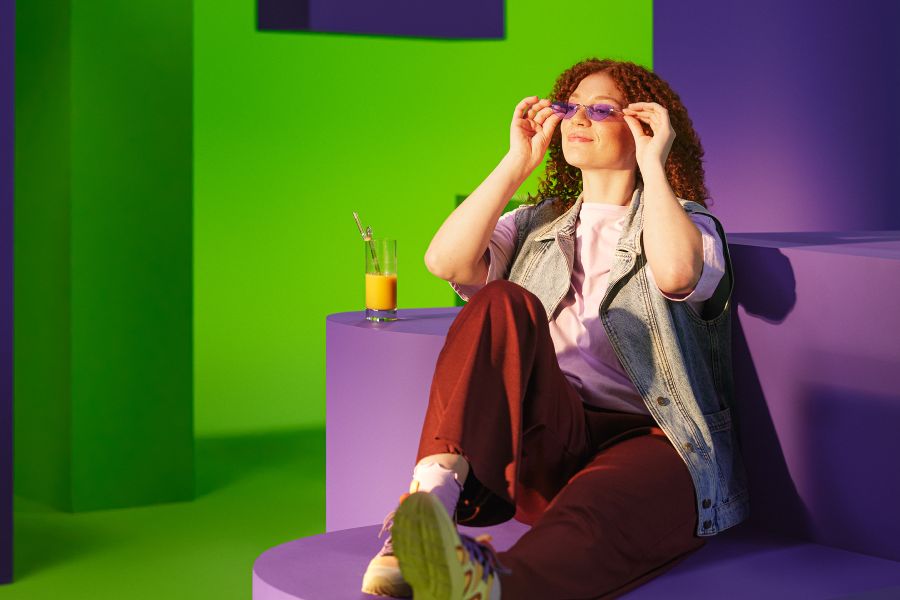 Vrouw met rode broek en zonnebril zit op paarse bank en glimlacht 